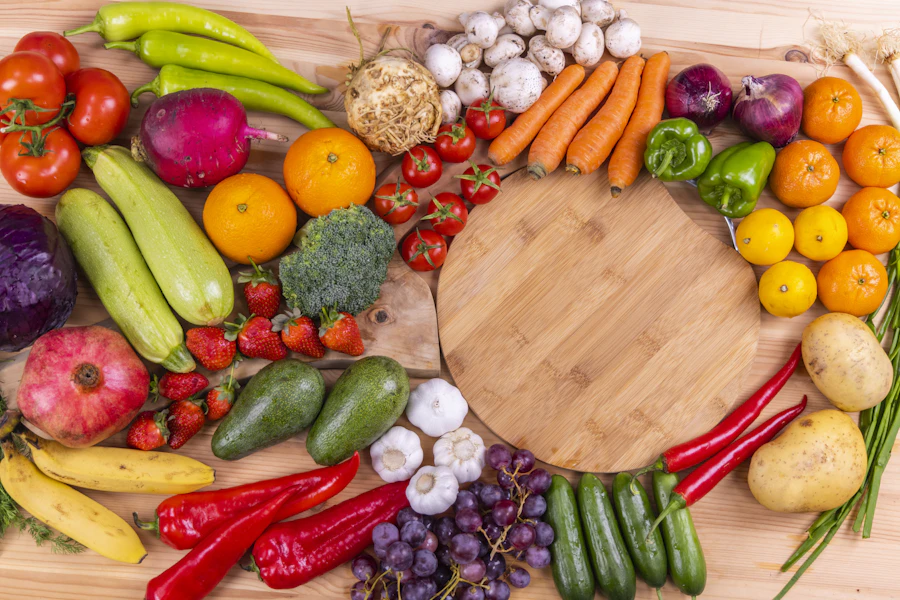 Legumes e frutas frescos, como tomate, cenoura, brócolis, pimentão e abobrinha, ao redor de uma tábua de madeira